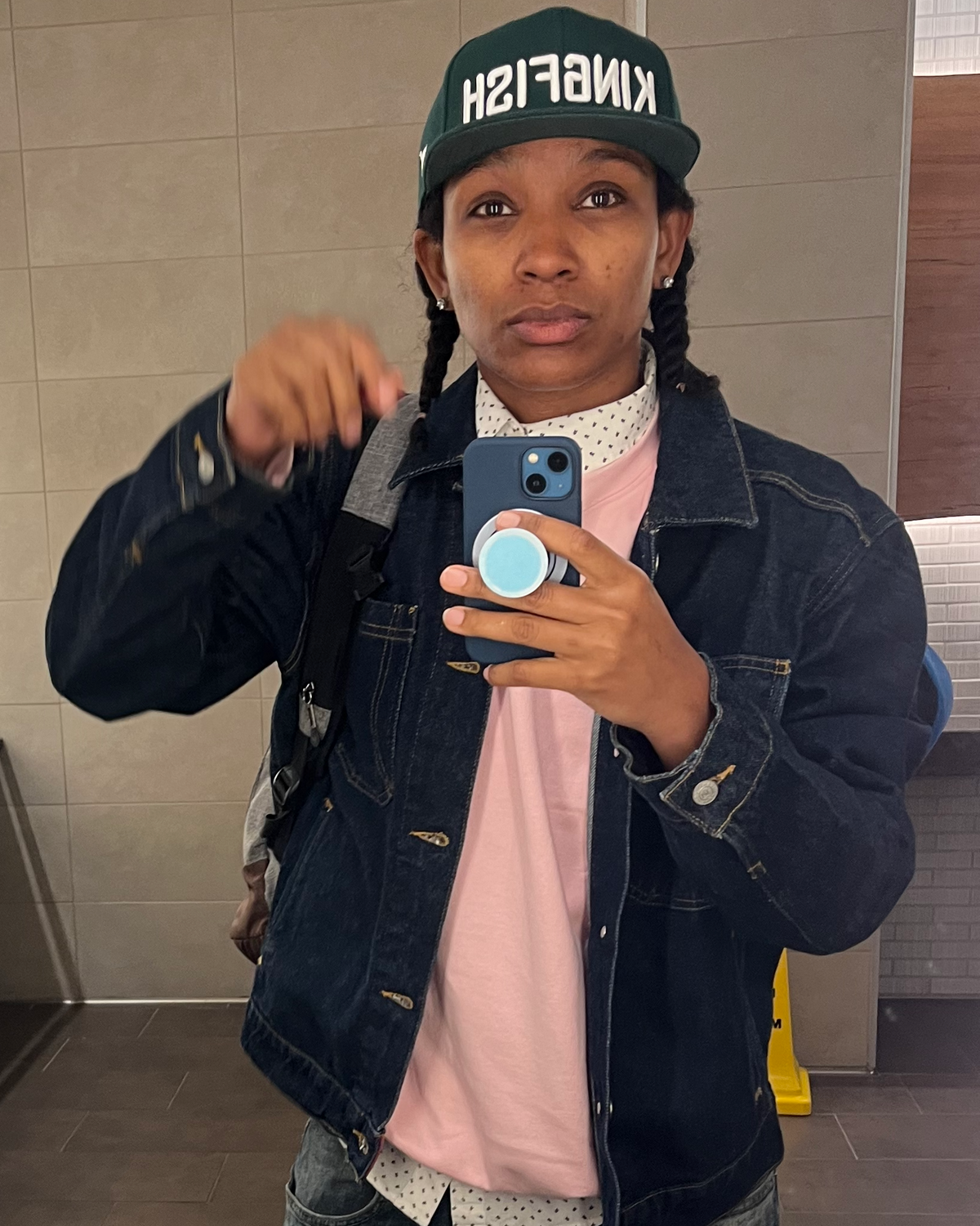 Person taking a mirror selfie wearing a denim jacket, pink shirt, and green cap indoors.